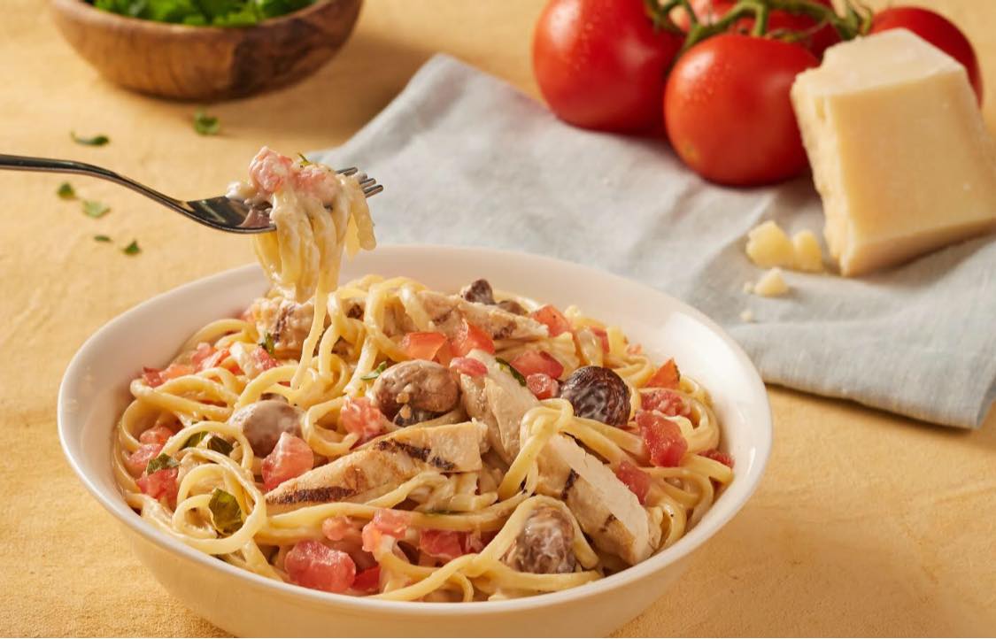 A large bowl of of chicken alfredo with mushrooms and tomato pieces. In the background of the photo there is a kitchen towel folded up with a block of parmesan and four on-the-vine tomatoes beside. In the top left corner of the photo there is a wooden bowl with salad in it.