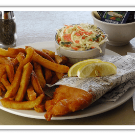 There is a large plate has a large piece of haddock fish and fresh and crisp french fries. There is two lemon slices on top of the battered and fried fish. On the back of the plate there is a glass serving dish with coleslaw.