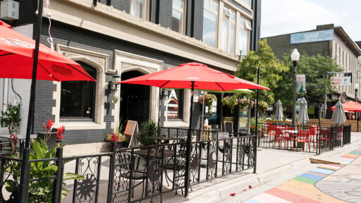 The picture consists of the front of the buidling with the outdoor patio. The red unbrellas are open and the black table and chairs are underneath each umbrella. In the bottom right corner of the photo there is colorful street art on the road.