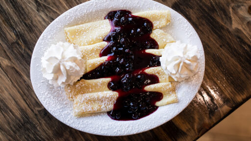 The is a large white plate in the center of the photo with 5 crepes folded up and placed all beside each other vertically. On top of the crepes is a blueberry sauce. On the right and left side of the plate, there is a swirl of whipped cream. Icing sugar is sprinkled over the entire dish.