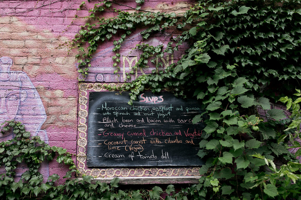 This is a photo of the side of the Soupcon Bistro building. In the center of the wall there is a chalk board with the days special soups. On the right hand side of the sign there is a vine plant growing up and covering the wall. On the left hand side of the sign there is art planted on the wall.