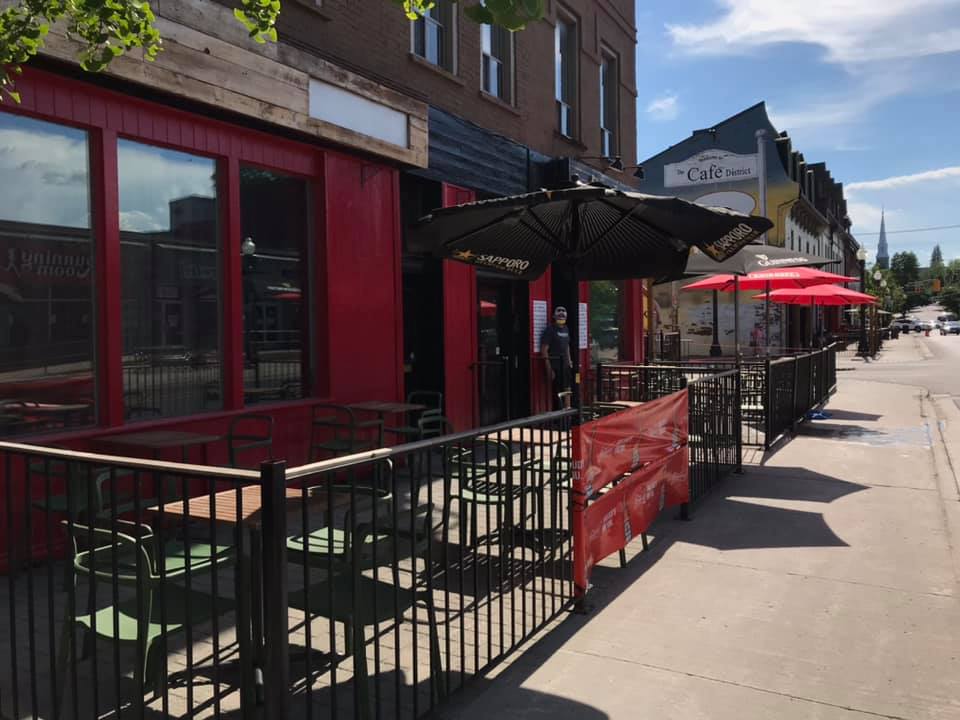 the photo consists of Spanky's outdoor patio. There is a black fence surrounding the patio. There are tables and picnic tables with umbrellas and some without. There is a Spanky's logo banner beside the entrance to the patio.