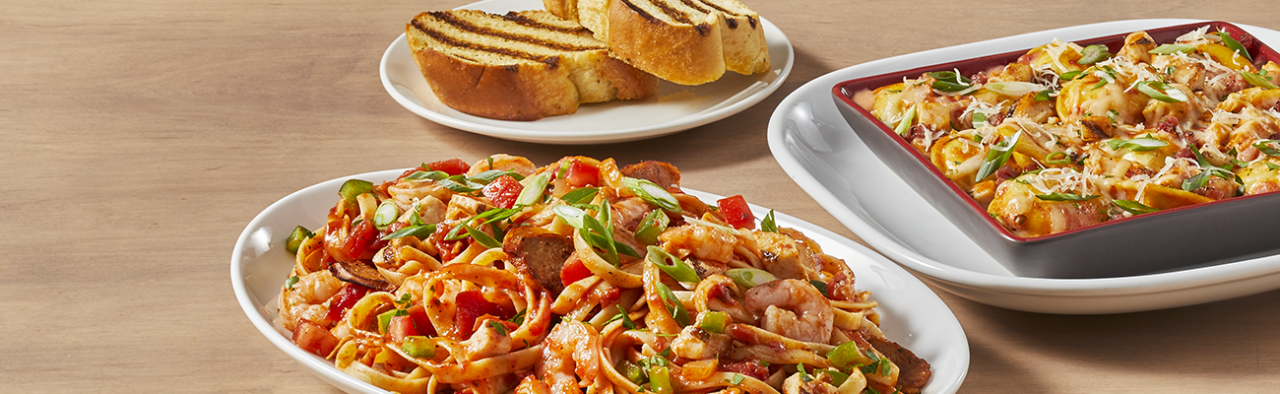 3 white plates placed in a circular shape. The plate at the top of the photo (the furthest plate) has two pieces of garlic bread with grill marks. The second plate on the right hand side of the photo has pasta in a bowl on the plate. The third plate is the closest plate and its located at he bottom of the photo. this plate takes up the majority of the photo and it is a pasta with spicy red pepper sauce, fettuccini noodles, shrimp, green onion, and spicy sausage.