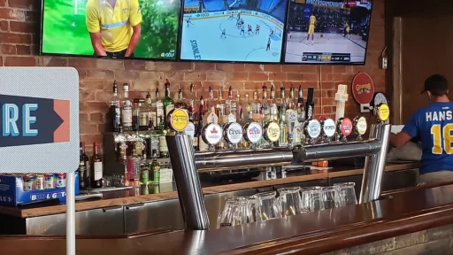 The photo consists of the bar at the restaurant and at the top of the photo there are 3 mounted flat screen tv's playing sports games. Underneath the tv's there are two shelves and across the shevles there is various different types of alcohol. At the bottom the the photo there is the bar countertop and behind that there are the beer taps.