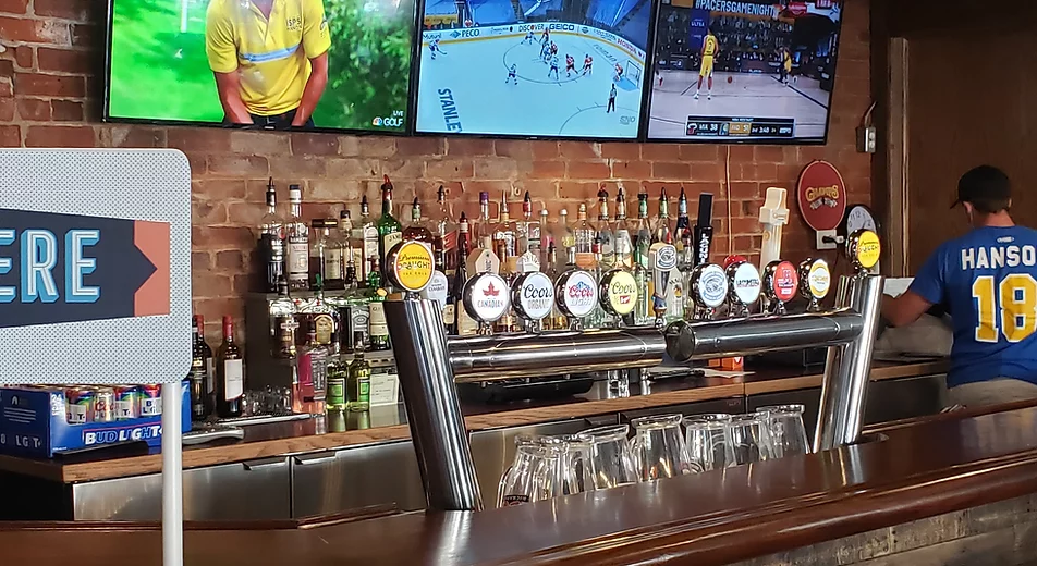 The photo consists of the bar at the restaurant and at the top of the photo there are 3 mounted flat screen tv's playing sports games. Underneath the tv's there are two shelves and across the shevles there is various different types of alcohol. At the bottom the the photo there is the bar countertop and behind that there are the beer taps.
