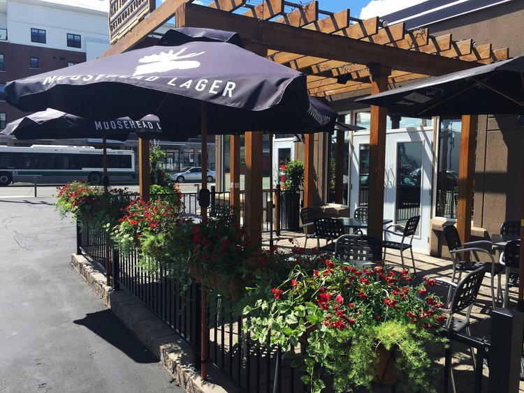 The photo consists of the outdoor patio of Kettle drums. There are black umbrellas over the tables and flower buckets hanging over the railings of the perimeter of the patio.