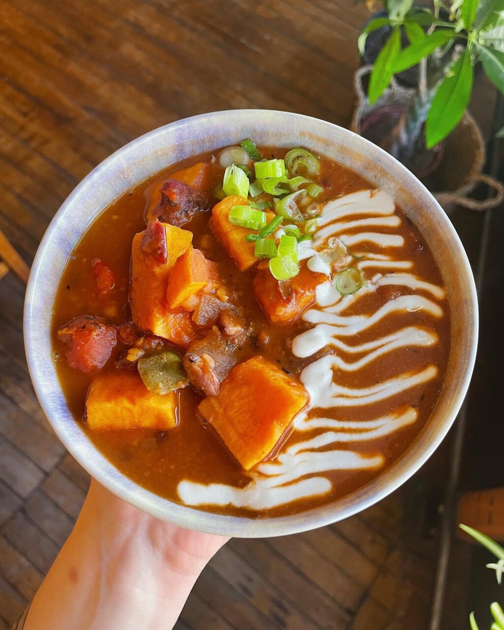 Hand holding a bowl of soup with chunks of sweet potato and white drizzled sauce