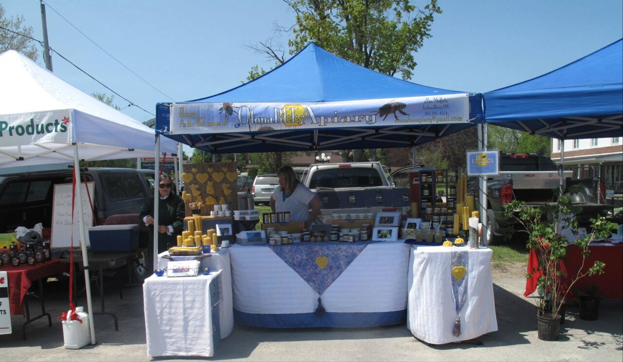 There is a vendor tent with the logo on it. Underneath the tent is a table with all the vendor product for sale on it. The vendor is selling candles, honey, and beeswax. There is the owner and woman selling the products behind the table under the tent. Beside the OtonaBEE Apiary is two other vendors on both side of the tent.