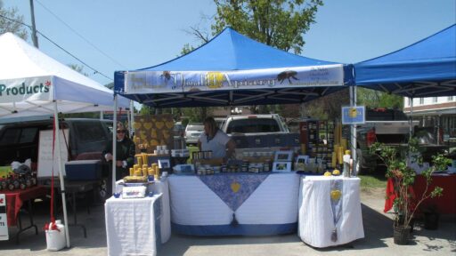 There is a vendor tent with the logo on it. Underneath the tent is a table with all the vendor product for sale on it. The vendor is selling candles, honey, and beeswax. There is the owner and woman selling the products behind the table under the tent. Beside the OtonaBEE Apiary is two other vendors on both side of the tent.