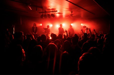 The photo is of a concert held in the Historic Red Dog Tavern. There are fans outlines cover the floor on the bottom half of the photo. There are only outlines of the people and their sihouettes filling the crowd. The three artists on stage are black outlines and silhouettes. In the background behind the artists is a red lights, making the photo just black and white.