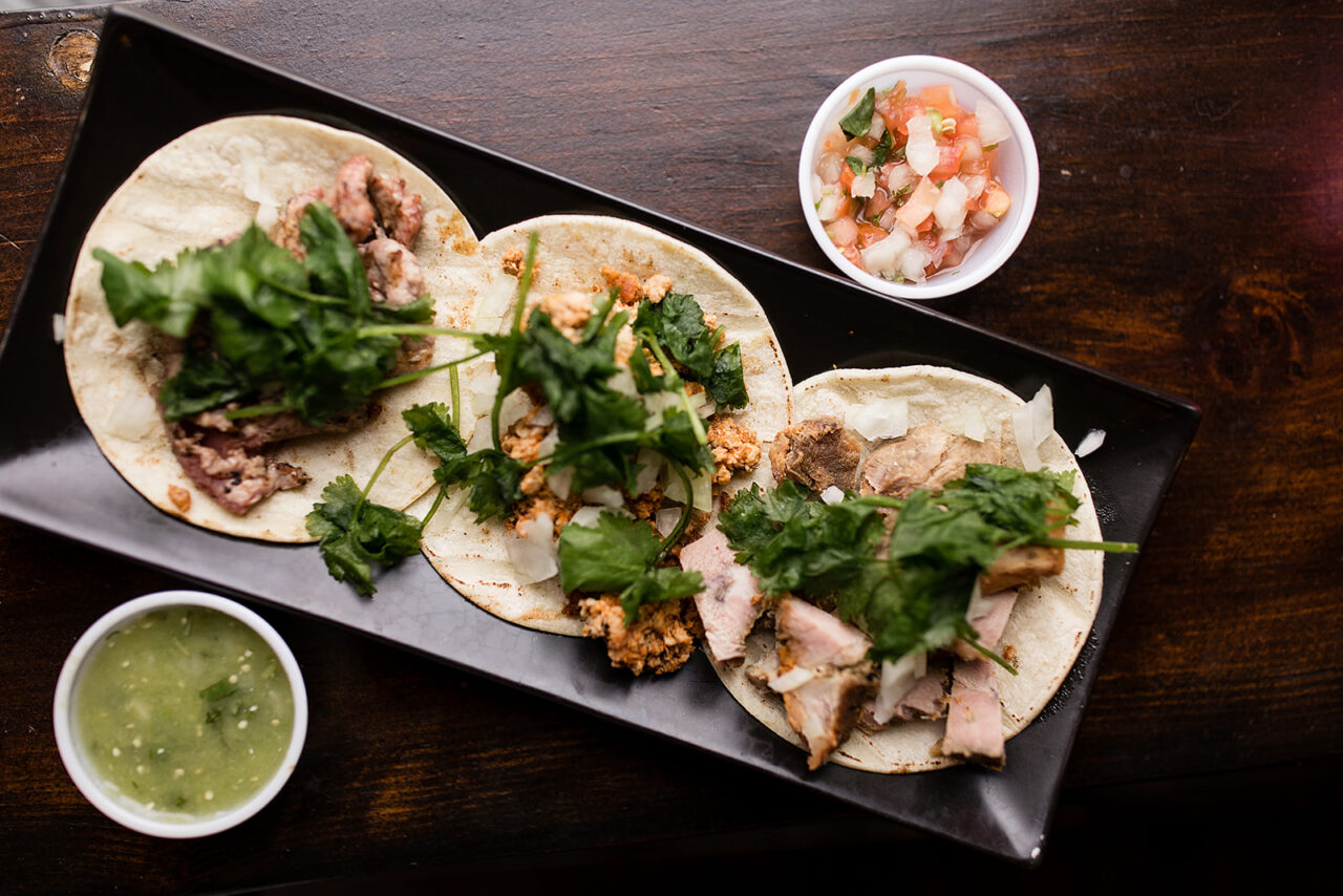 There is a rectangle black plate diagonally through the photo. On the plate there are three different types of tacos, beef, pork, and chicken. All have cilantro, onion, and are on a white tortilla. On the top right hand side of the photo there is a salsa in a serving cup and on the botton left corner there is a green sauce.