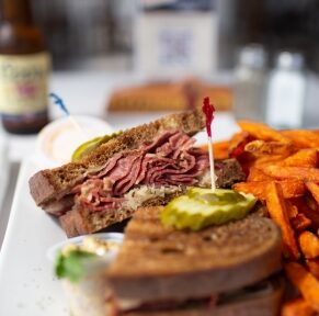 There is a large plate with a rueben snadwich on toasted whole wheat bread. The sandwich is cut in half and there are sliced pickle on top of the sandwiches, held in place with sanwich picks. Beside the sanwich is sweet potato fries.