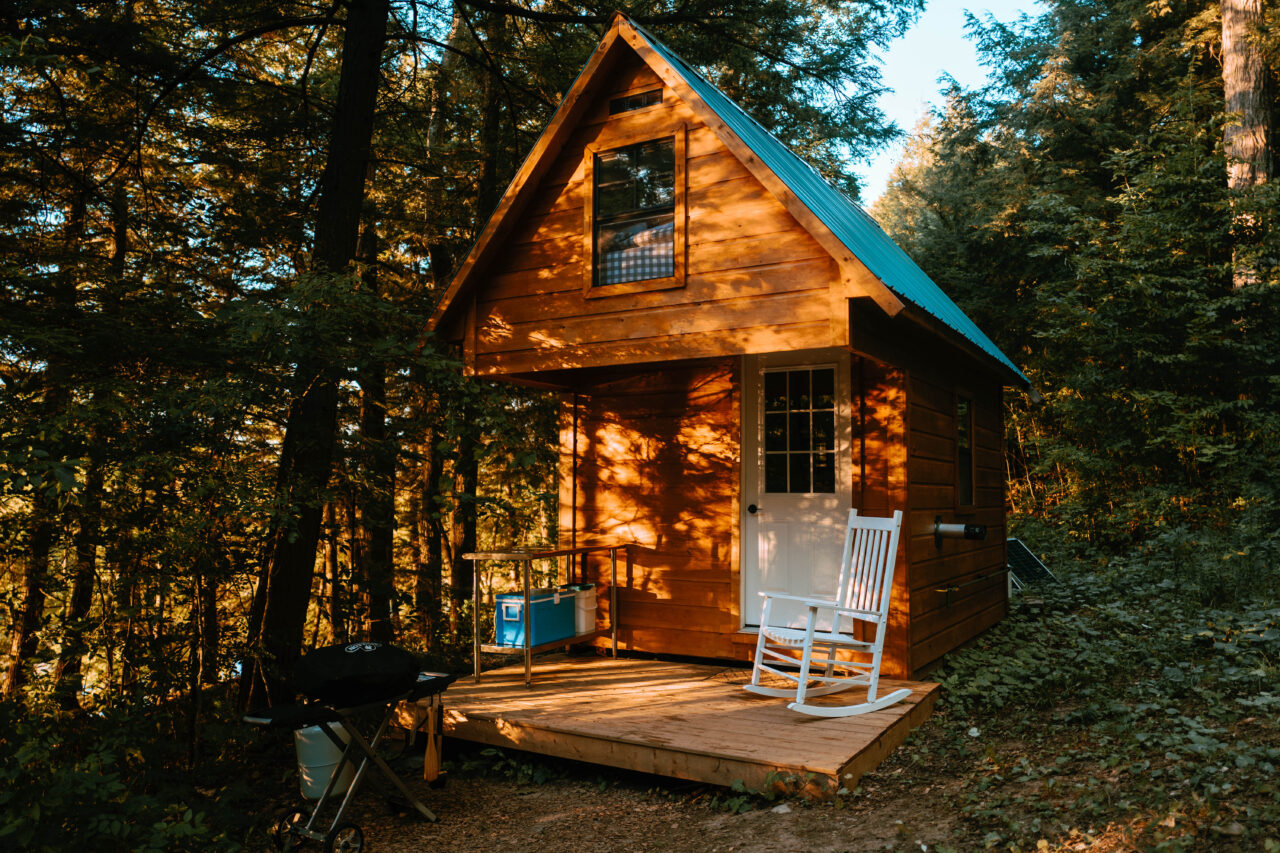 Wood bunkie with green roof in the woods with a small front porch and white rocking chair