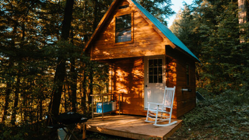 Wood bunkie with green roof in the woods with a small front porch and white rocking chair