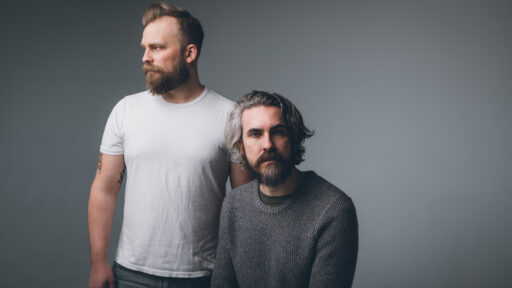 two men standing in front of a grey background. One looking at the camera and one looking away