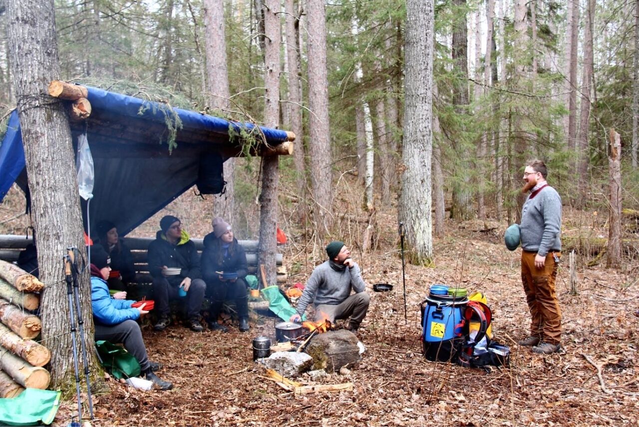 A 3 walled log shelter in the woods  in the fall with 4 people sitting, a man kneeling by a fire cooking and a man standing talking to the group