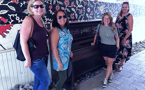 four women standing in front of a piano against a wall with a floral motif