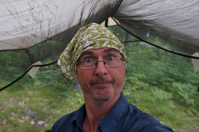 A person wearing a bandana on their head stands under a tent in a forest