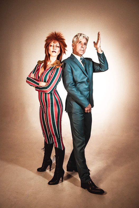 a man and a woman standing back to back in front of a brown backdrop, dressed like David Bowie