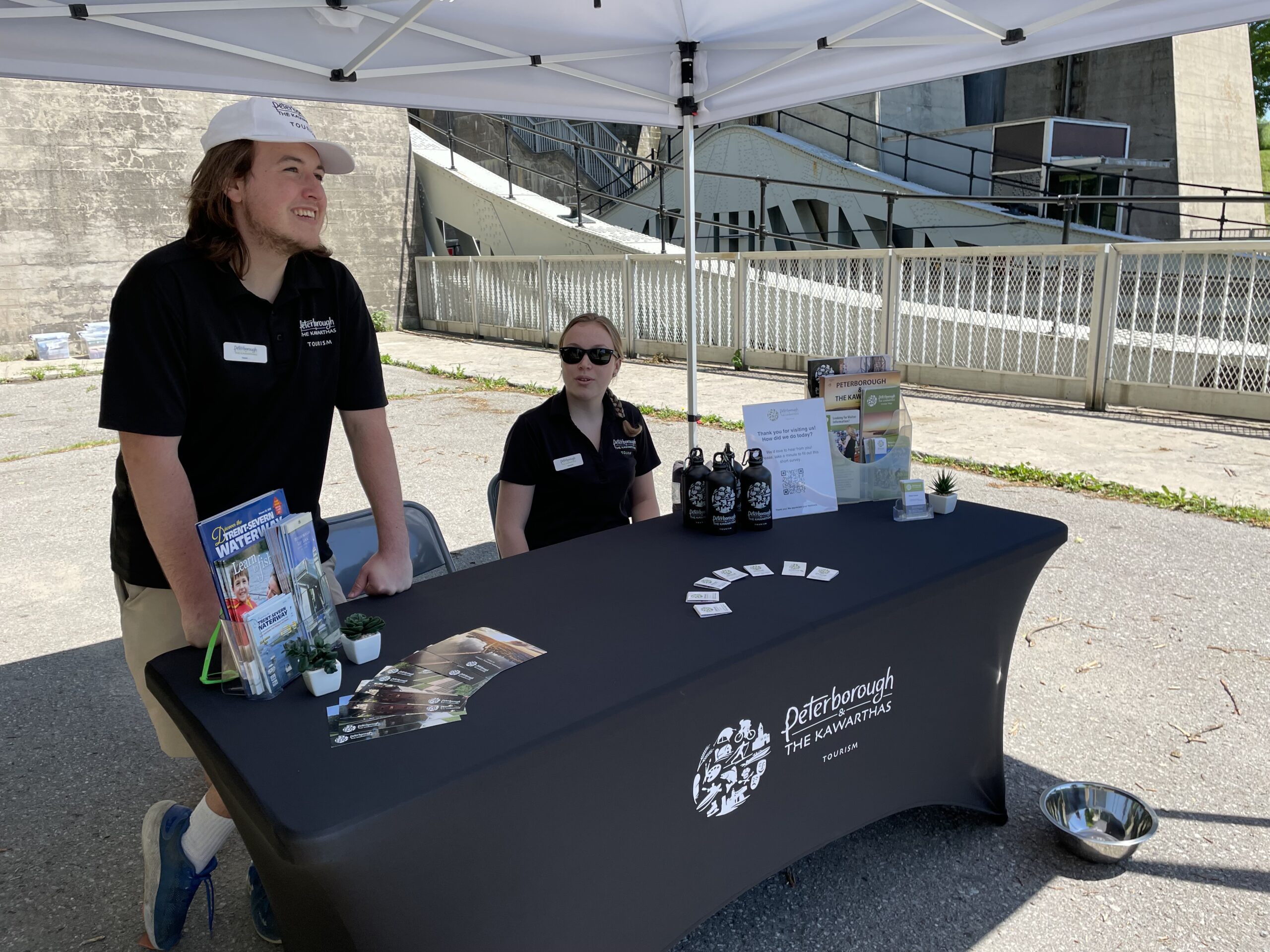 Person in black shirt, white hat standing behind table with brochures and black table cloth. A person in a black shirt and sunglasses sitting behind the table sitting
