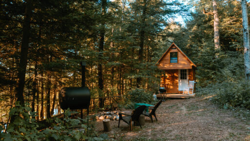 Bunkie in the woods with small front porch with a white rocking chair and smoker beside a fire pit