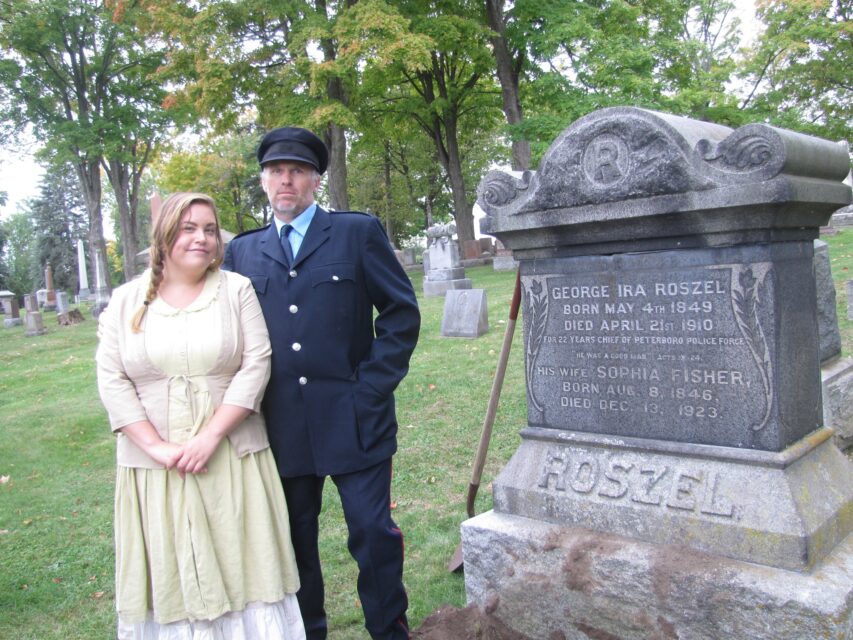 a man and a women dressed in early settelers costumes standing by a gravestone