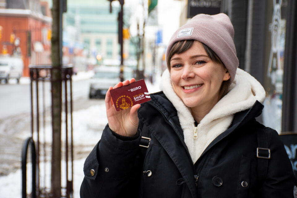 Picture of a women in a winter coat and toque standing on a sidewalk in Downtown Peterborough in the winter. She is smiling and holding a BORO gift card.