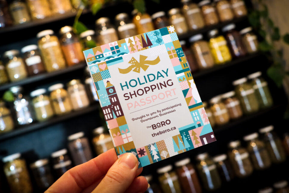 picture of someone's hand holding a rectangular card that says "Holiday Shopping Passport" on the front. The card is in front of a background of jars on a shelf