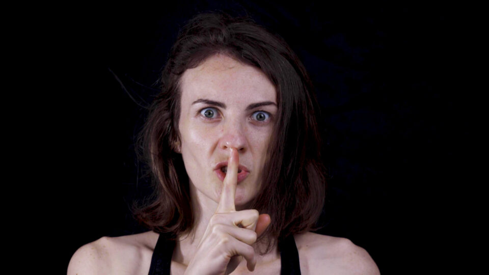 Woman with dark hair holding index finger over lips indicating "shh"