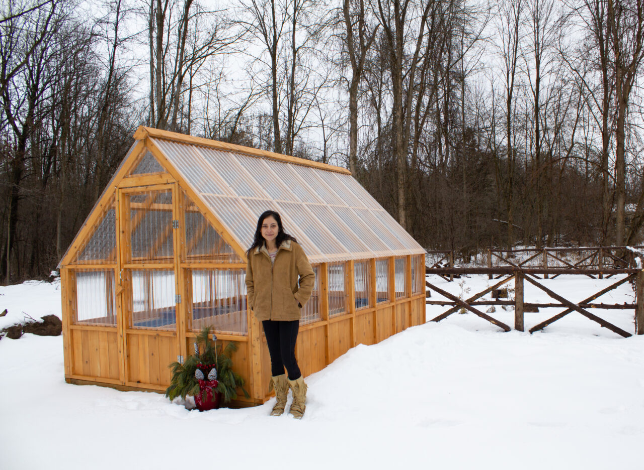 woman in brown jacket standing outside in winter, in front of wooden greenhouse structure