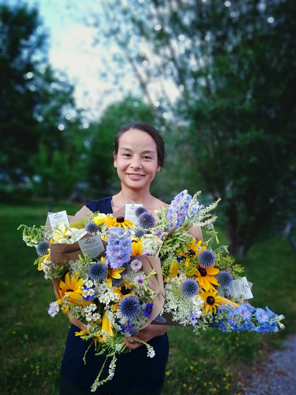 Women smiling with bouquet of flowers in front of body. Green trees in background