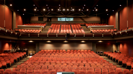 an image of an empty theatre from the stage. the seats are red, and span across 2 levels