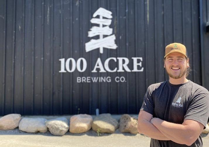 100 Acre Brewing Company owner Bentley Vass (young white man with baseball cap and black shirt) is standing with his arms crossed in front of the Brewery - a blue building with a white 100 Acre Brewing Co sign on the front