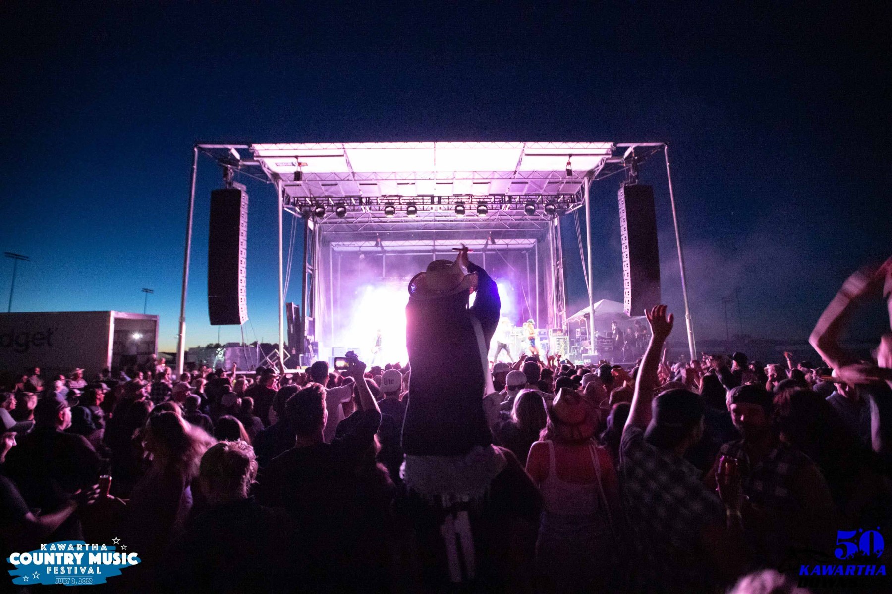 a photo of a crowd at an outdoor concert at night. featured in the center is a person propped up on someone's shoulders