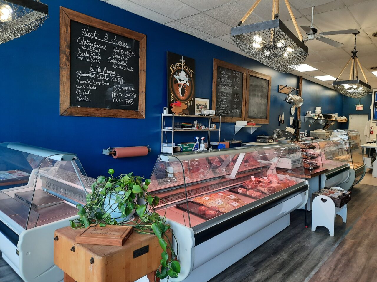 Butcher shop counter full of product in front of blue wall with chalk board sign menus