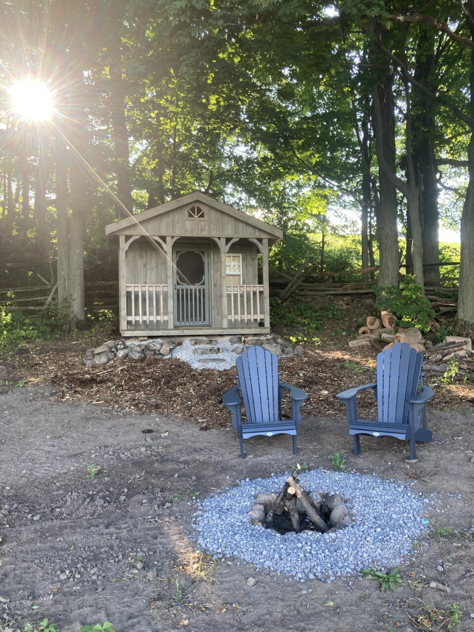 Wood bunkie with small front porch and two chairs in front of fire pit with forest in background