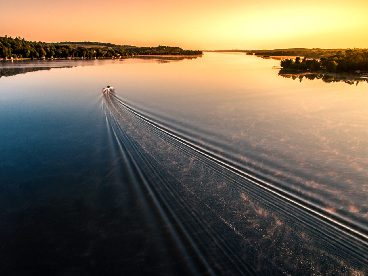 Aerial view looking towards a body of water with ripples from a boat in the distance travelling towards an orange sky with the sun setting