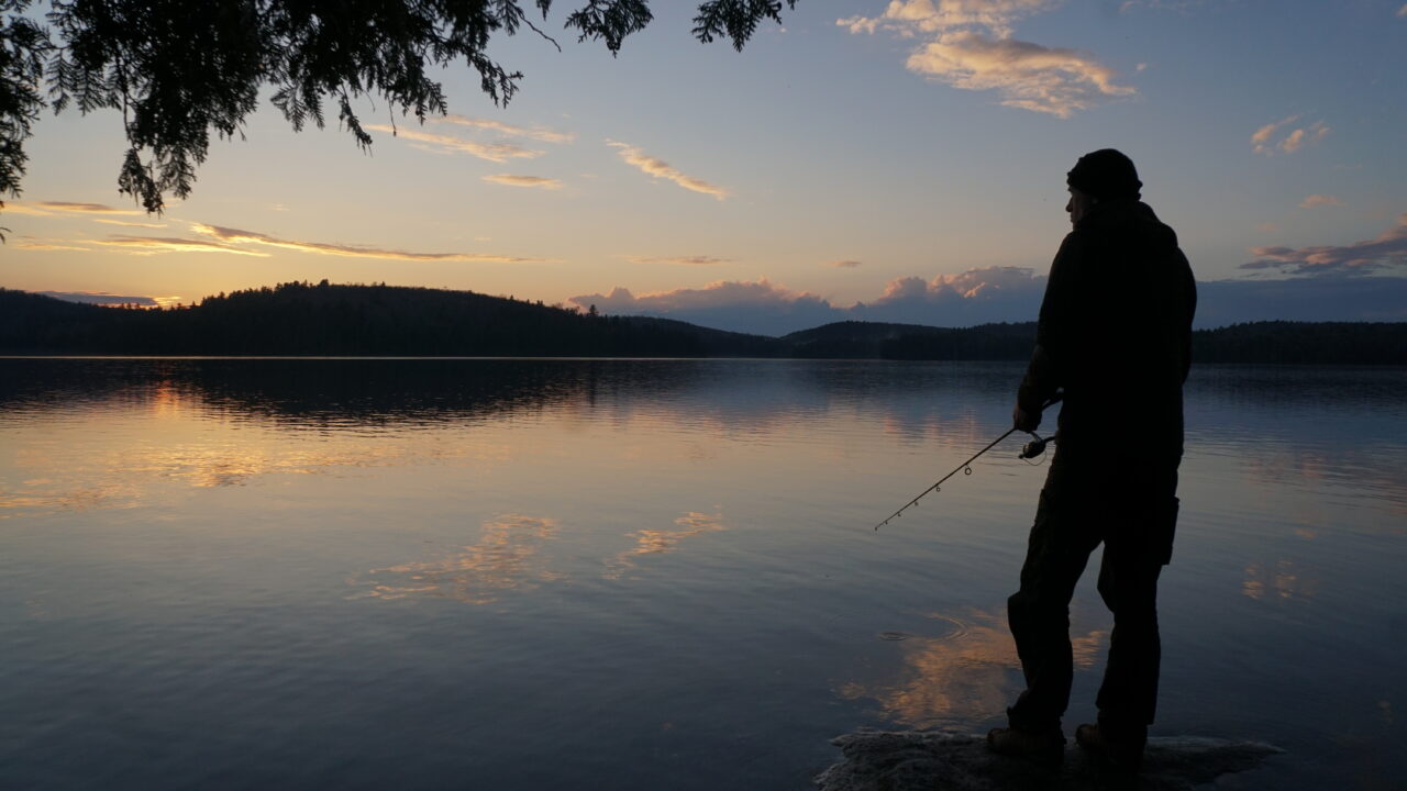 a person fishing at dusk, on the shore of a lake