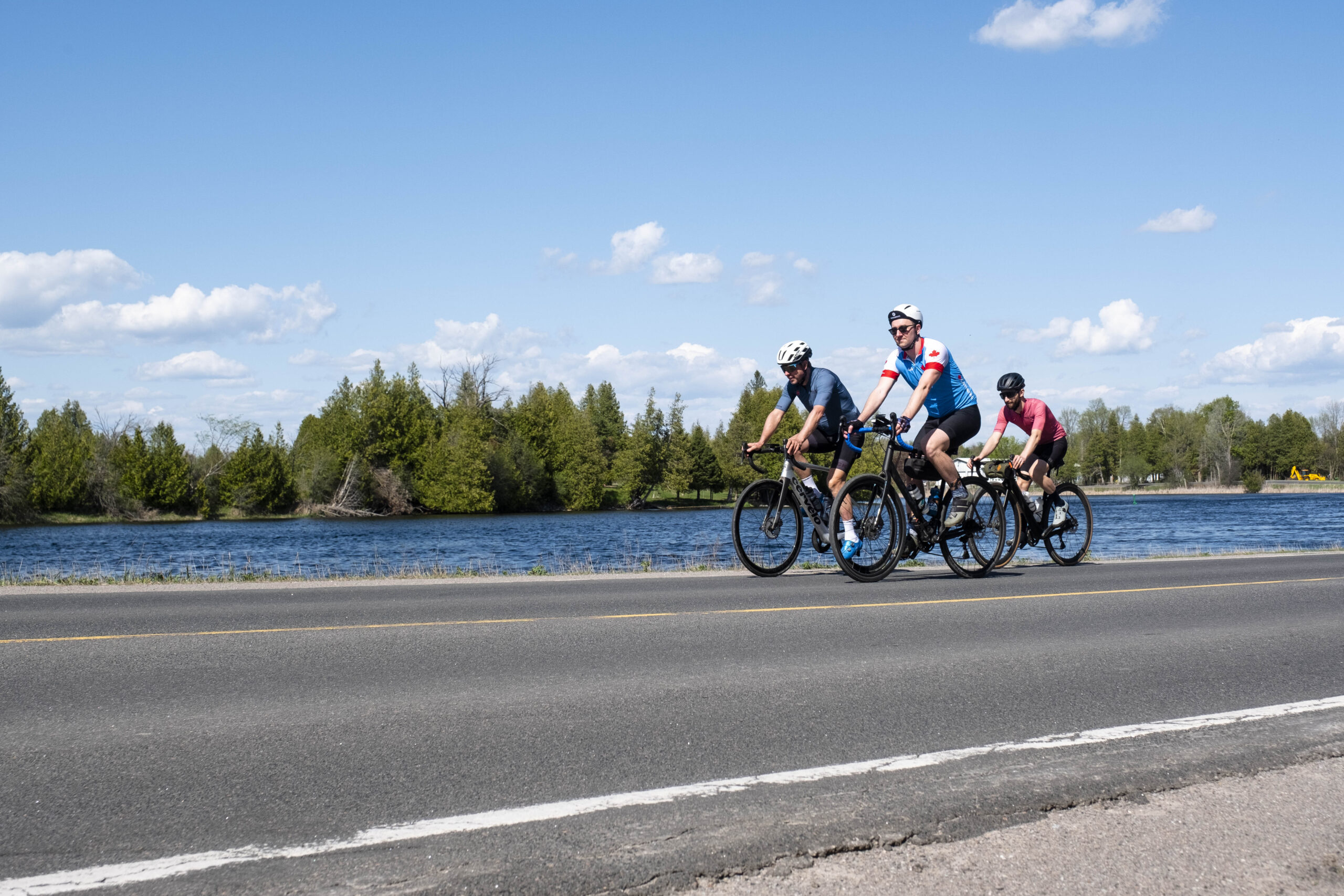 three cyclists riding down a road in Peterborough & the Kawarthas, in front of a lake