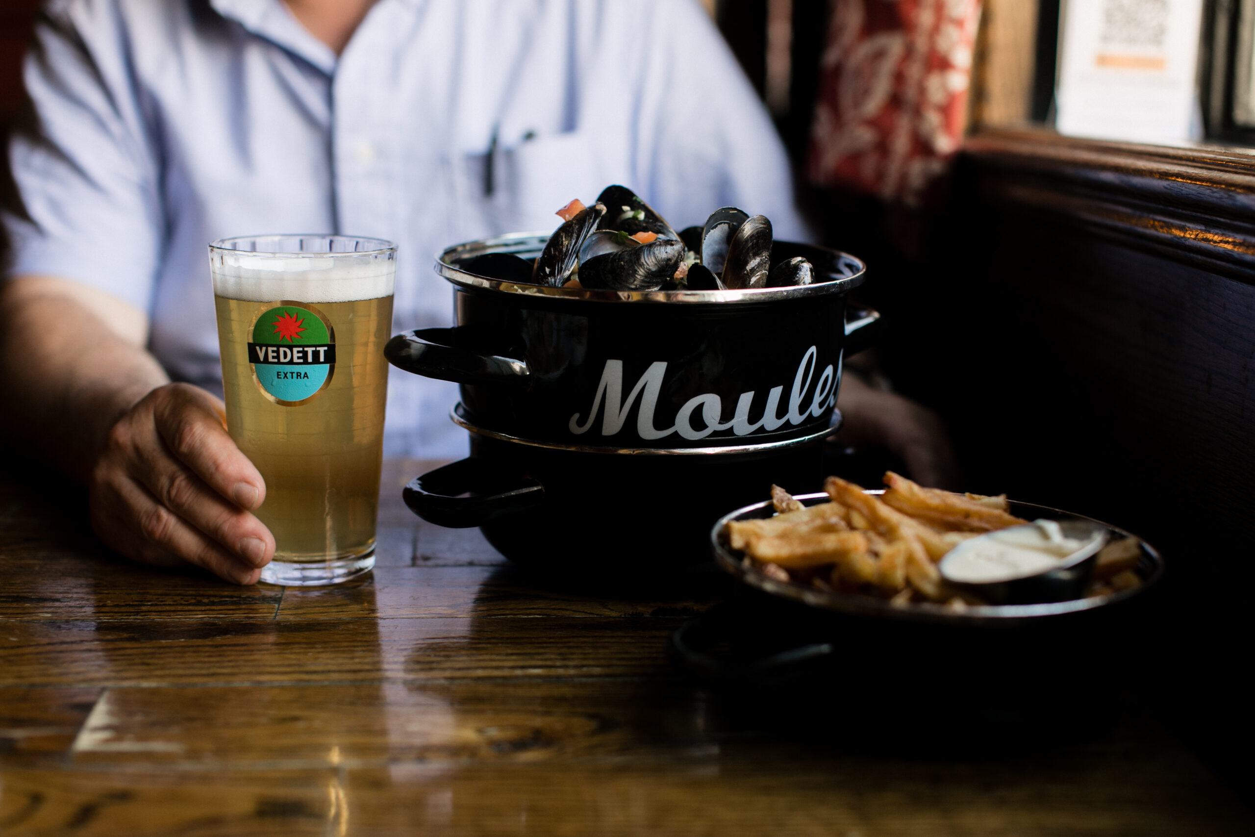 A table with pots of mussels and frites; a person's hand holding a glass of beer with their body blurred in the background.