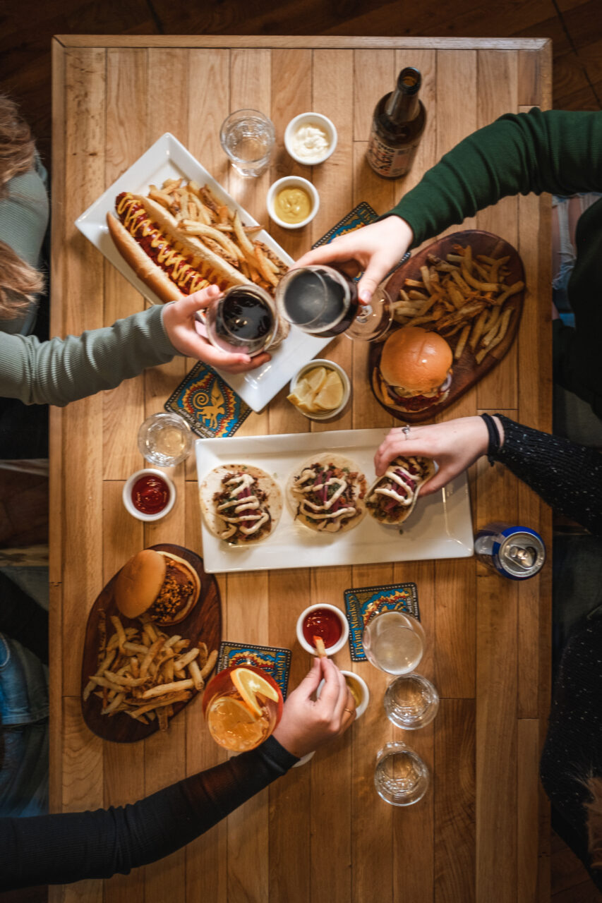 Point-of-view looking down at a table filled with a variety of Dirty Burger menu items and drinks with four hands reaching over the table, two cheers-ing and two picking up food.