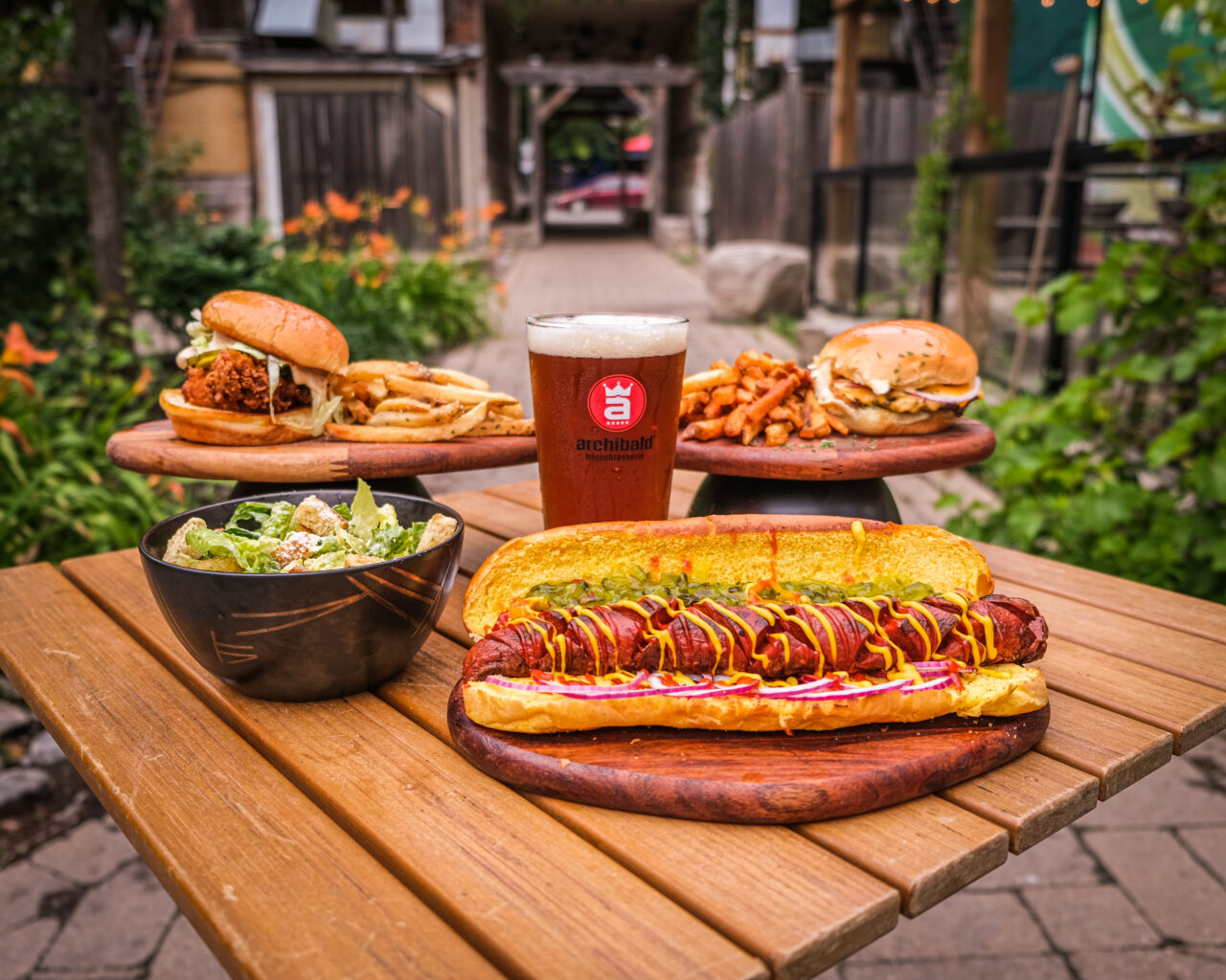Food and a beer sitting on a wood table on the Dirty Burger patio surrounded by greenery.
