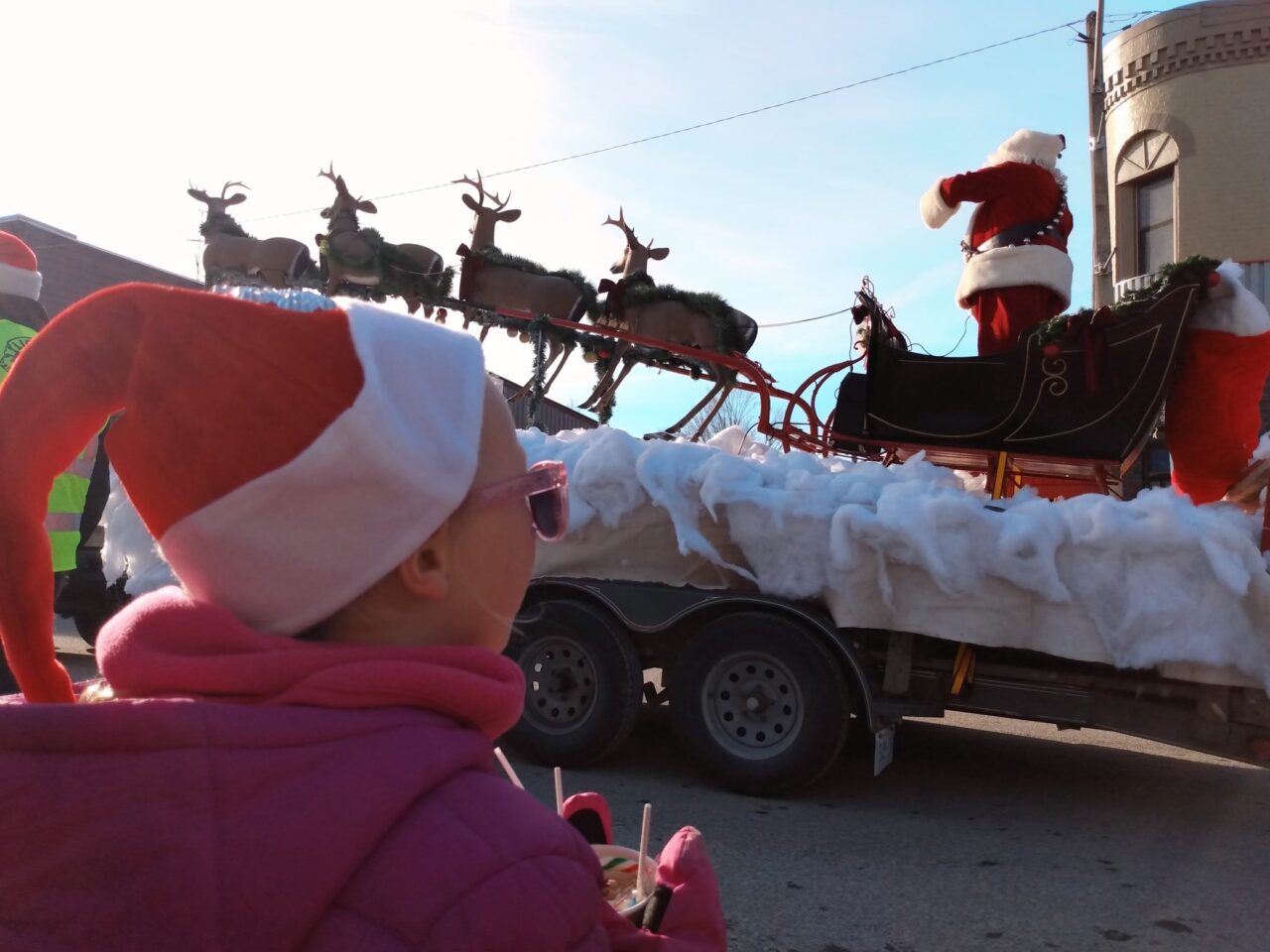 Profile photo of a child in a santa hat with sun glasses looks up at Santa's arrival at the Lakefield Santa Claus parade.
