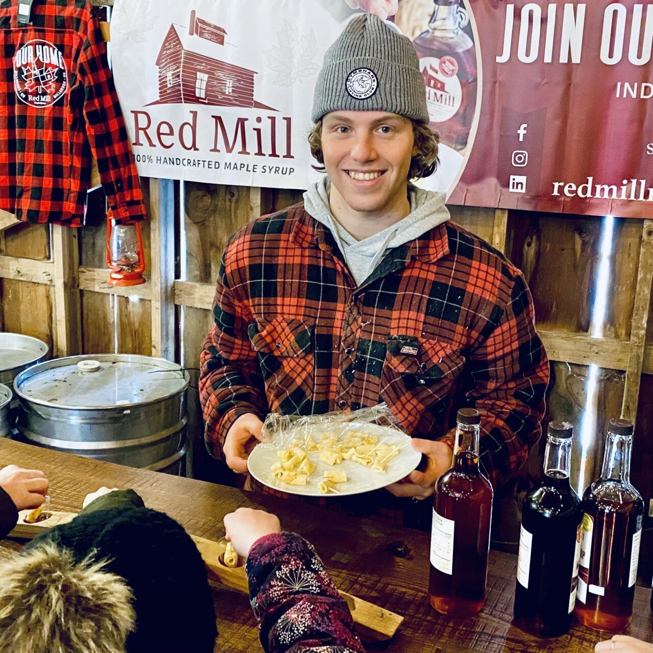Jeremy from Red Mill Maple in red plaid jack and grey toque holding a plate of maple candy behind the maple tasting bar