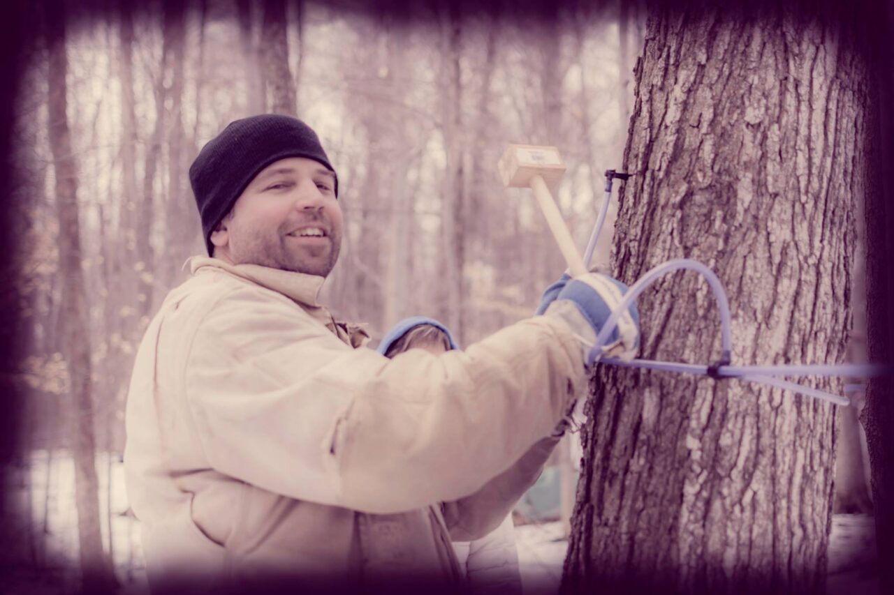 A man smiling at the camera while using their hands to tap a tree